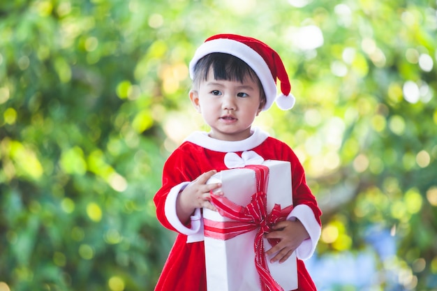 Nettes asiatisches Baby, das Sankt-Anzug in der Hand hält schöne Geschenkbox trägt