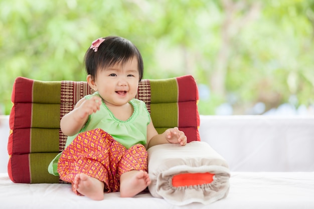 Nettes asiatisches Baby im thailändischen traditon Kleid, das mit dem Kissen lächelt mit Glück sitzt