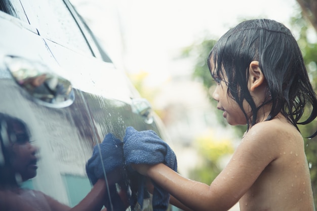 Nettes asiatisches Kind, das am Sommertag ein Auto mit Schlauch wäscht