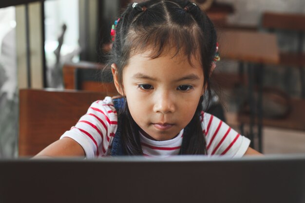 Nettes asiatisches Kindermädchen, das auf Laptop im Café mit Spaß und Glück verwendet und spielt