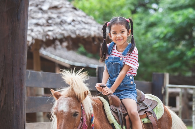 Nettes asiatisches Kindermädchen, das ein Pony im Bauernhof mit Spaß reitet