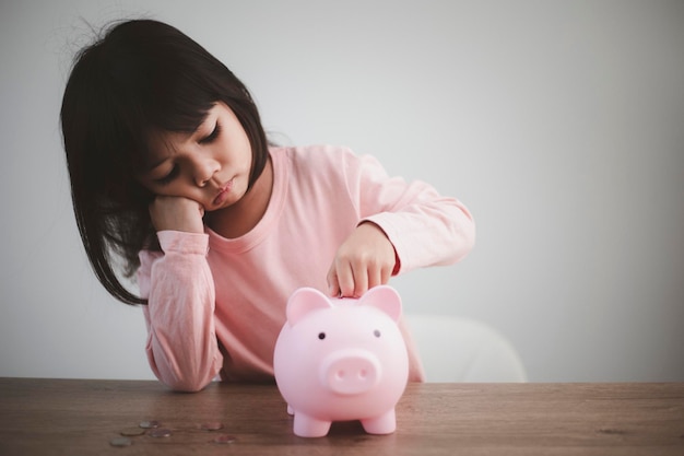Nettes asiatisches Kindermädchen, das Geld in das Sparschwein steckt, um Geld für die Zukunft zu sparen
