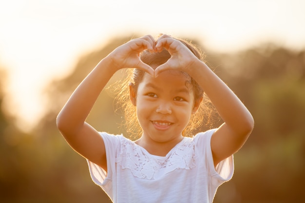 Nettes asiatisches Kindermädchen, das Herzform mit Händen im Feld mit Sonnenlicht macht