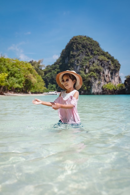 Nettes asiatisches Kindermädchen, das Hut und Sonnenbrille trägt, die Spaß haben, im Meer zu spielen.