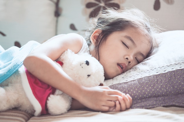 Nettes asiatisches Kindermädchen, das ihren Teddybären im Bett schläft und umarmt