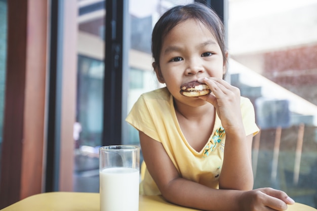 Nettes asiatisches Kindermädchen, das Milch trinkt und Keks in der Frühstückszeit isst