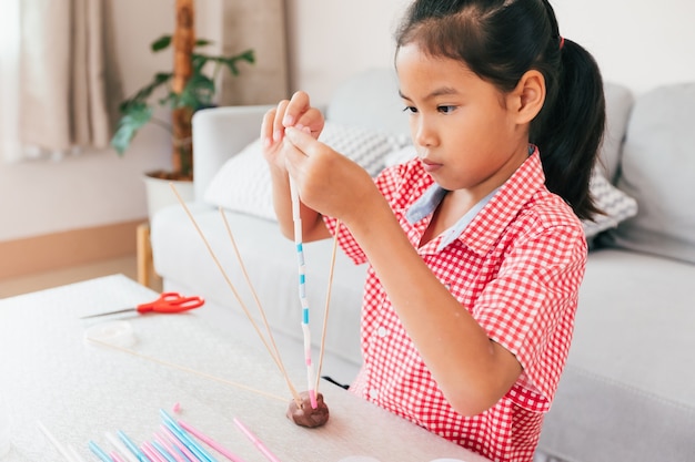 Nettes asiatisches Kindermädchen, das mit Knetmasse und Strohhalmen spielt und schafft. Kind konzentrierte sich mit Knetmasse, die ein Molekülmodell baute. Kind und Handwerk mit recyceln Dingen.