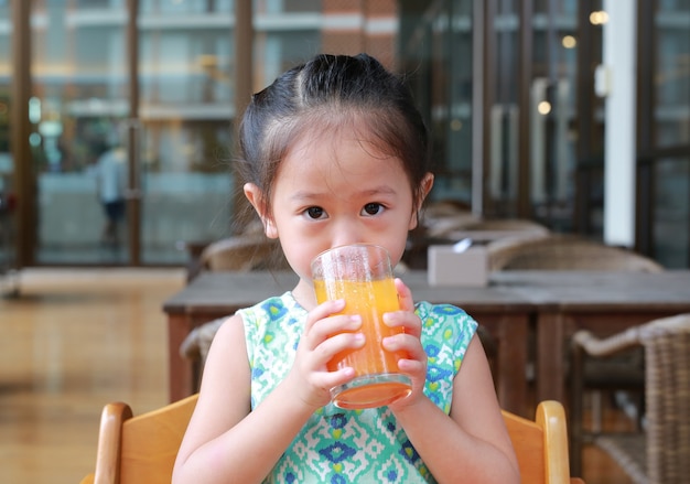 Nettes asiatisches Kindermädchen, das Orangensaft am Restaurant mit dem Schauen der Kamera trinkt.