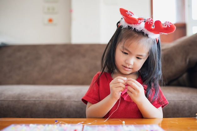 Nettes asiatisches Kindermädchen, das Weihnachtskostüm trägt, das Perlen auf eine Schnur mit Absicht und Spaß zu Hause einfädelt.