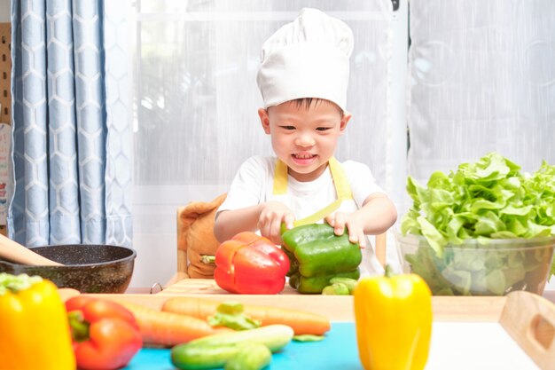 Nettes asiatisches kleines Jungenkind, das Kochmütze und Schürze trägt, die Spaß beim Vorbereiten haben, gesundes Essen in der Küche kochend, lustige Innenaktivitäten für Kindergartenkinder