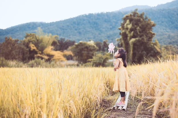 Nettes asiatisches kleines Kindermädchen, das mit Windkraftanlage auf dem Reisfeld im Weinlesefarbton spielt und durchbrennt