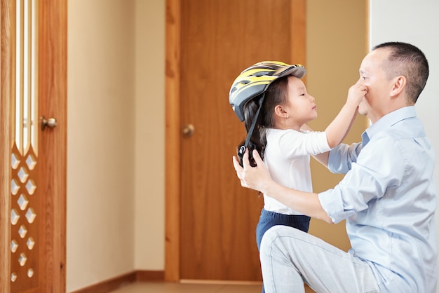 Nettes asiatisches kleines Mädchen, das Gesicht ihres Vaters berührt, der ihren Fahrradhelm aufsetzt, bevor er Wohnung verlässt