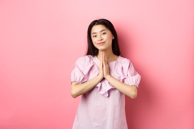 Nettes asiatisches Mädchen sagt Danke, lächelt und sieht glücklich aus, zeigt namaste Geste in Dankbarkeit, im Kleid gegen rosa Hintergrund stehend.