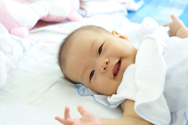 Nettes asiatisches Säuglingsbaby im weißen Kleid liegt und lächelt auf ihrem Bett.