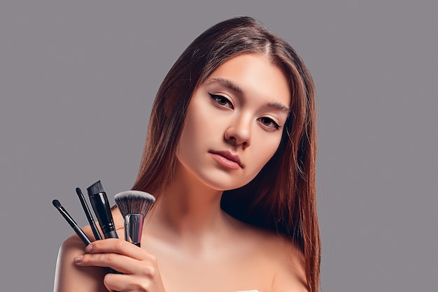Nettes attraktives Mädchen, das Make-upbürsten lokalisiert auf grauem Hintergrund hält. Hautpflegekonzept. Spa-Behandlungen, Kosmetik, Make-up.