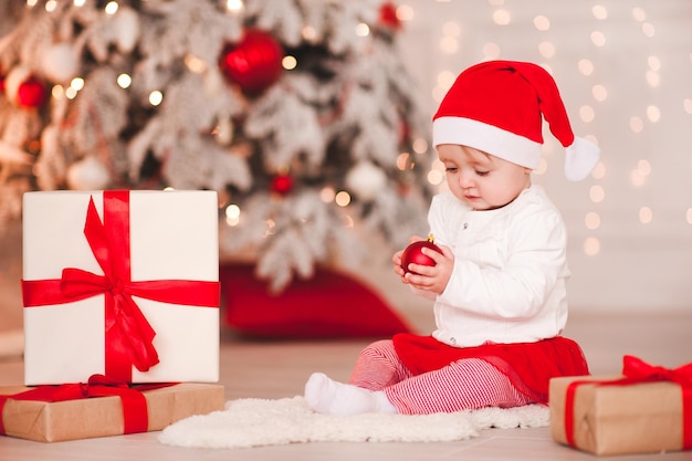 Nettes Baby 1 Einjahres, das Sankt-Hut trägt, der über Weihnachtshintergrund aufwirft. Sitzen auf dem Boden mit Weihnachtskugel. Ferienzeit.