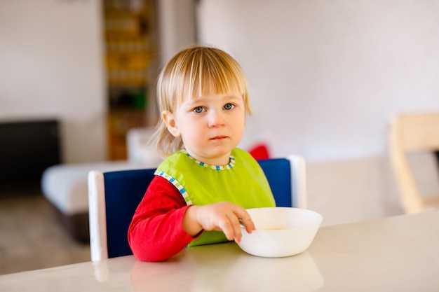 Nettes Baby, das auf hohem Kinderstuhl sitzt und Gemüse allein in der weißen Küche isst.