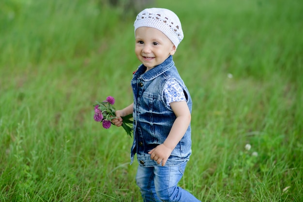 Nettes Baby, das im schönen Park mit bunter Blume auf Sommersaison geht, die er lächelt und glücklich stehenden Naturhintergrund im Freien steht.