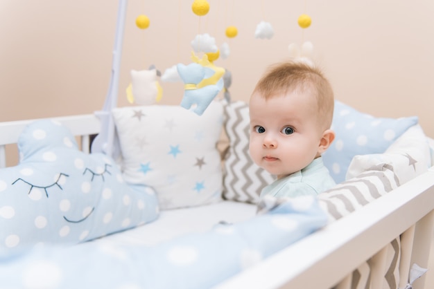 Nettes Baby, das in einem weißen runden Bett sitzt. Leichter Kindergarten für kleine Kinder. Spielzeug für Kinderbett. Lächelndes Kind, das mit Handy des Filzes im sonnigen Schlafzimmer spielt.