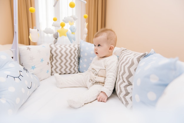 Nettes Baby, das in einem weißen runden Bett sitzt. Leichter Kindergarten für kleine Kinder. Spielzeug für Kinderbett. Lächelndes Kind, das mit Handy des Filzes im sonnigen Schlafzimmer spielt.