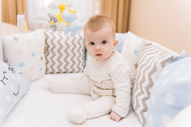 Nettes Baby, das in einem weißen runden Bett sitzt. Leichter Kindergarten für kleine Kinder. Spielzeug für Kinderbett. Lächelndes Kind, das mit Handy des Filzes im sonnigen Schlafzimmer spielt.