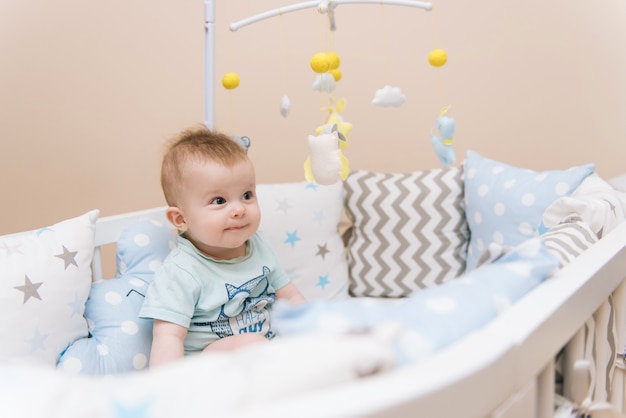 Nettes Baby, das in einem weißen runden Bett sitzt. Leichter Kindergarten für kleine Kinder. Spielzeug für Kinderbett. Lächelndes Kind, das mit Handy des Filzes im sonnigen Schlafzimmer spielt.