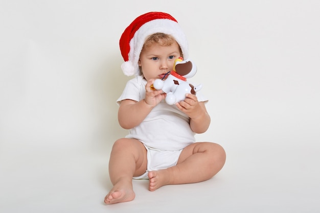 Nettes Baby im Weihnachtsmannhut und im Bodysuit, die auf Boden mit Plastikhund in Händen sitzen, Säugling, der mit seinem Spielzeug spielt