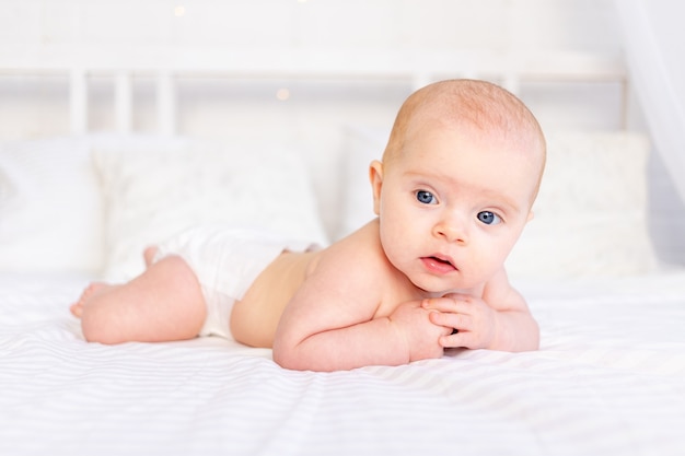 Nettes Baby in einer Krippe auf einem weißen Baumwollbett, das auf dem Bauch im Kinderzimmer liegt und lächelt, neugeborener Morgen