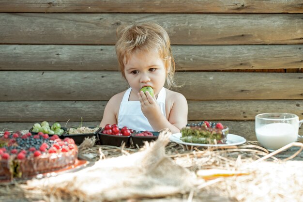 Nettes Baby isst grünen Apfel am Tisch mit Beeren-Himbeer-Stachelbeer-Kirsch-Johannisbeer-Obstkuchen und Tasse Milch im Freien auf Holz