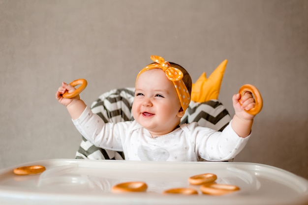 Nettes Baby lächelt, das in einem Babystuhl sitzt und Bagels isst