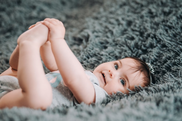 Nettes Baby mit blauen Augen, die auf grauem Blatt im Schlafzimmer liegen und ihre Beine halten