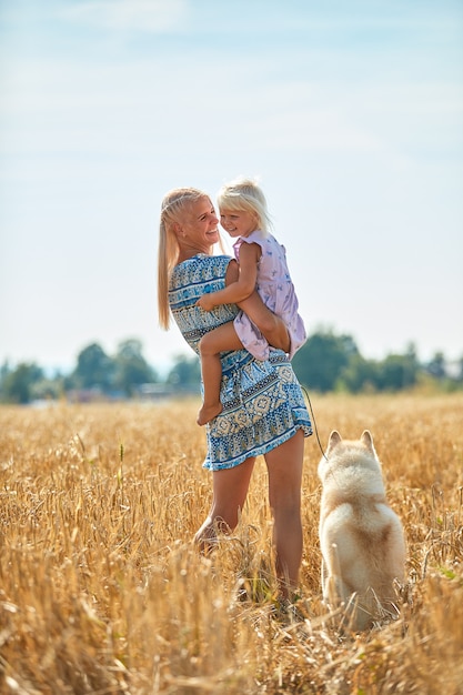 Nettes Baby mit Mutter und Hund auf Weizenfeld. Glückliche junge Familie genießt die gemeinsame Zeit in der Natur. Mama, kleines Baby und Hundehusky, die draußen ruhen. zusammengehörigkeit, liebe, glückskonzept.