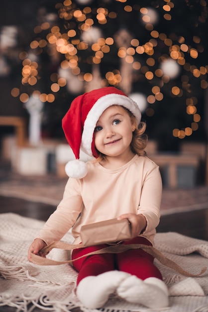 Nettes blondes Kindmädchen geöffnete Weihnachtsgeschenkbox, die unter Weihnachtsbaum sitzt