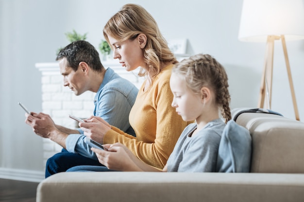 Nettes ernstes blondes kleines Mädchen, das ihr Telefon hält und anschaut, während es ordentlich ihre Eltern sitzt und sie auch ihre Telefone benutzen