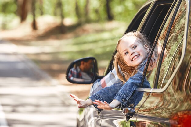 Nettes fröhliches kleines kaukasisches Mädchen, das im Auto sitzt und aus dem Fenster schaut und lächelt