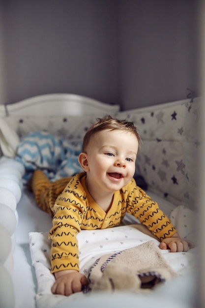 Nettes fröhliches lächelndes blondes Kleinkind mit schönen blauen Augen, die auf dem Bauch in seinem Kinderbett am Morgen liegen und aufwachen.