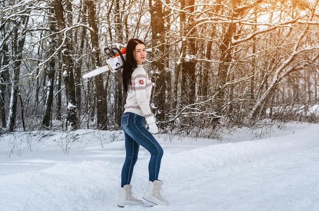 Nettes fröhliches Mädchen geht mit einer Kettensäge durch den Schnee im Wald