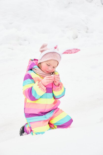 Nettes fröhliches Vorschulmädchen mit Caranal-Häschenohren, das im Schneeschnee im April Osterhasen-Winterspaß spielt