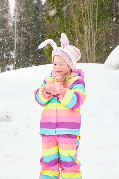 Nettes fröhliches Vorschulmädchen mit Caranal-Häschenohren, das im Schneeschnee im April Osterhasen-Winterspaß spielt