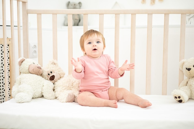 Nettes gesundes Mädchen, das auf einem Babybett mit Teddybären auf weißer Bettwäsche im Schlafzimmer zu Hause sitzt Baby schläft in ihrer Krippe