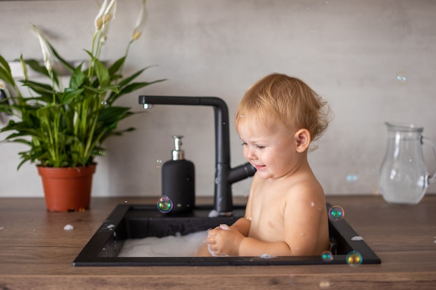 Nettes glückliches Baby mit dem Spielen mit Wasser und Schaum in einer Küchenspüle zu Hause