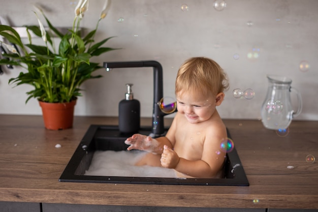 Nettes glückliches Baby mit dem Spielen mit Wasser und Schaum in einer Küchenspüle zu Hause