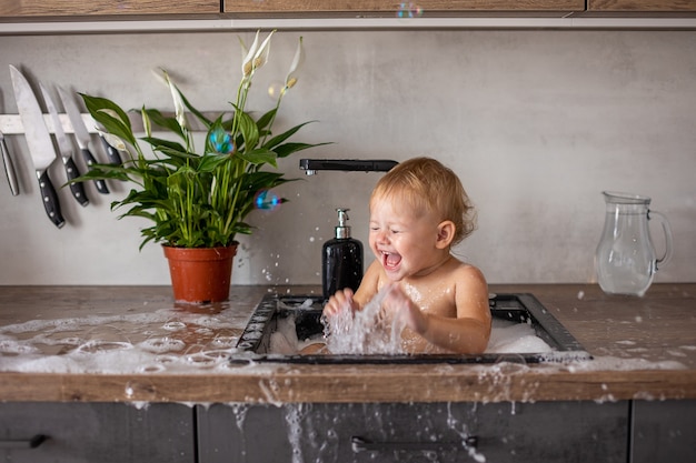 Nettes glückliches Baby mit dem Spielen mit Wasser und Schaum in einer Küchenspüle zu Hause