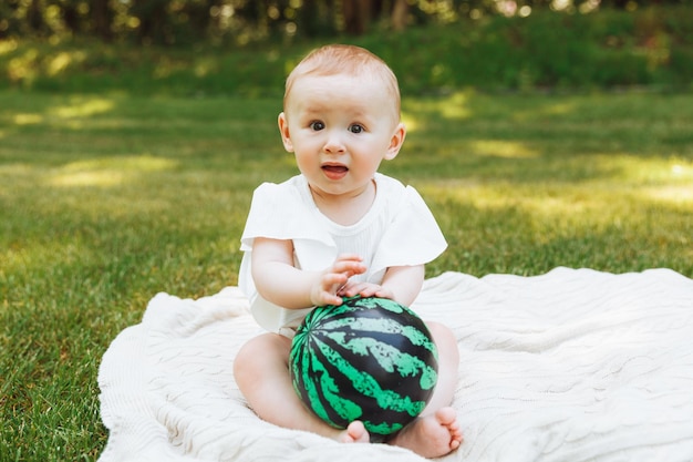 Nettes glückliches Babymädchen mit einem Ball sitzt auf grünem Gras im Park