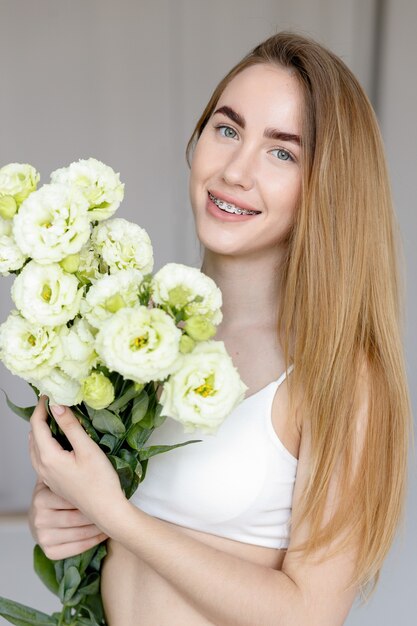 Nettes glückliches junges Mädchen im weißen leeren T-Shirt, das einen Blumenstrauß hält und die Blumen betrachtet, den Geruch genießend. schönes blondes jugendlich Mädchen mit Zahnspange an den Zähnen lächelnd