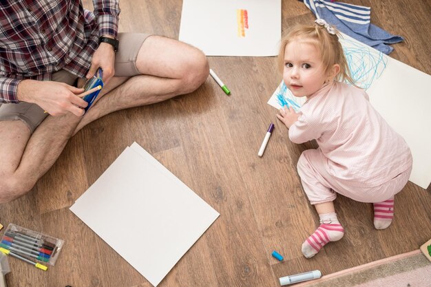 Nettes glückliches Kind zeichnet mit farbigen Markern auf weißem Papier, das neben seinem Vater auf dem Boden sitzt