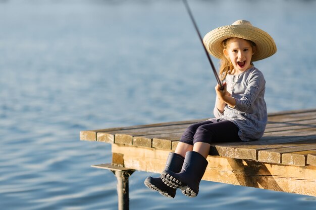 Nettes glückliches kleines Kindermädchen in Gummistiefeln, die vom hölzernen Pier auf einem See fischen.