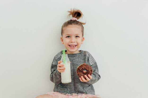 Nettes glückliches kleines Mädchen mit lustiger Frisur mit Schokoladendonut und einer Flasche Milch