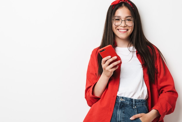 Nettes glückliches Teenager-Mädchen, das lässiges Outfit trägt, das isoliert über weißer Wand steht und Handy hält