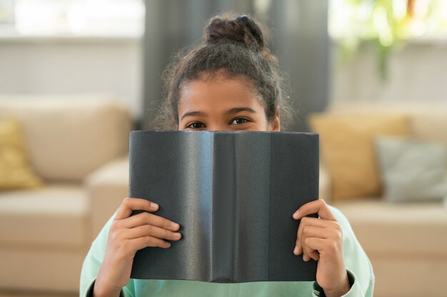 Nettes Grundschulmädchen, das vor der Kamera im Wohnzimmer gegen die Couch mit Kissen sitzt und dich aus dem offenen Buch anspäht